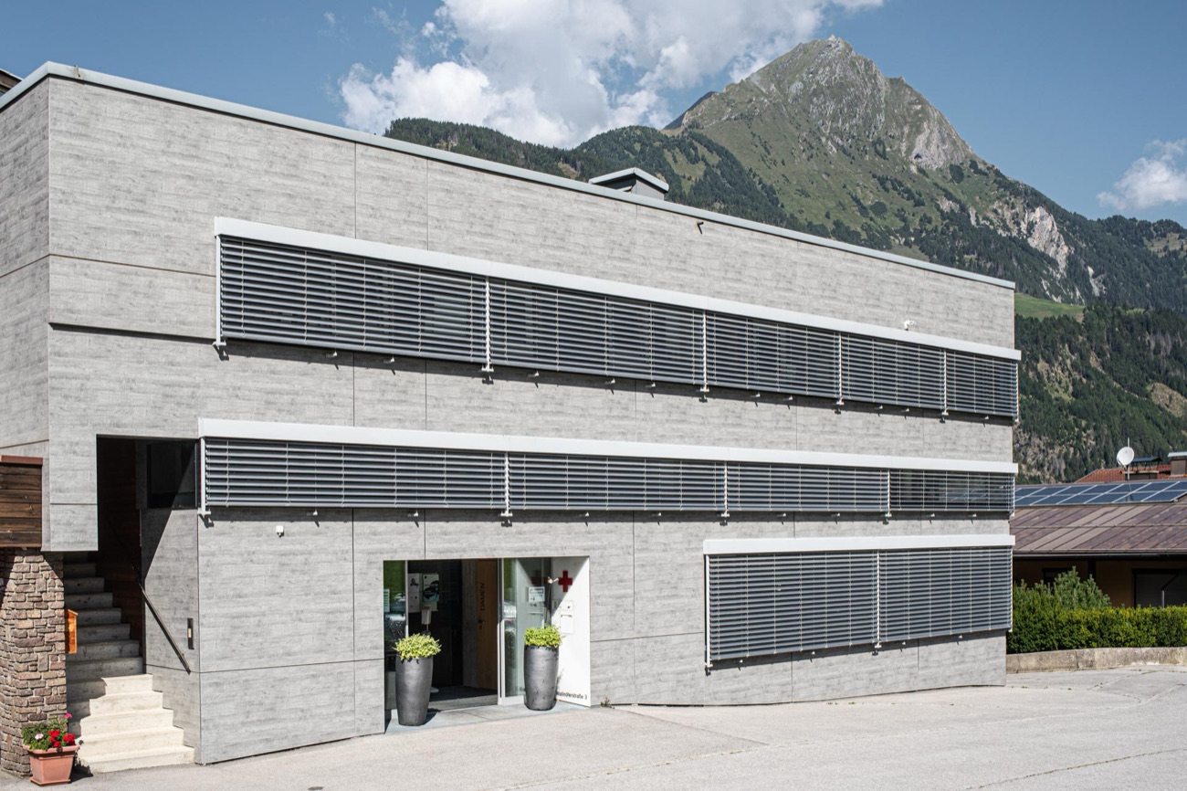 Eingang der Ordination mit rotem Kreuz an der Fassade, im Hintergrund die Berge rund um Matrei in Osttirol.