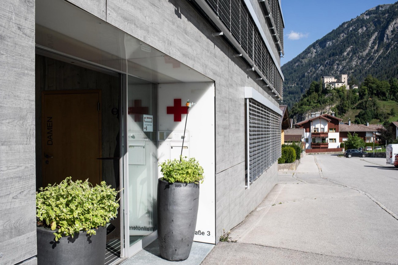 Eingang der Ordination mit rotem Kreuz an der Fassade, im Hintergrund die Berge rund um Matrei in Osttirol.